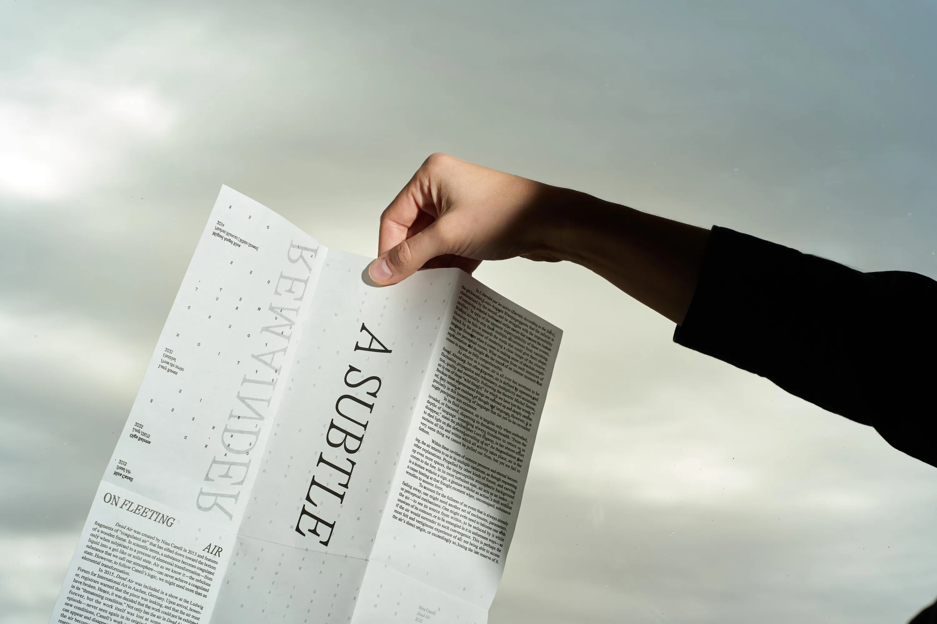 Hand holding the unfolded A Subtle Remainder broadsheet against a cloudy sky