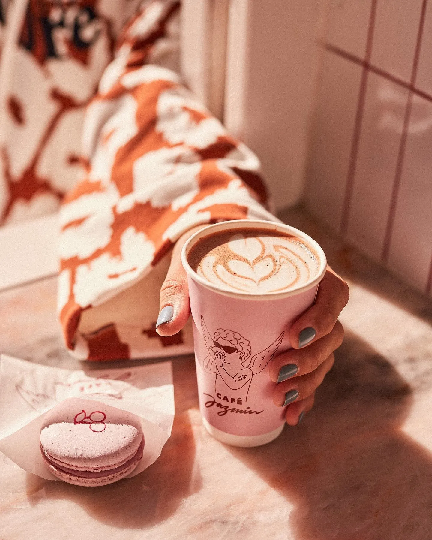 Hand holding a pink Café Jazmín cup of latte beside a macaron