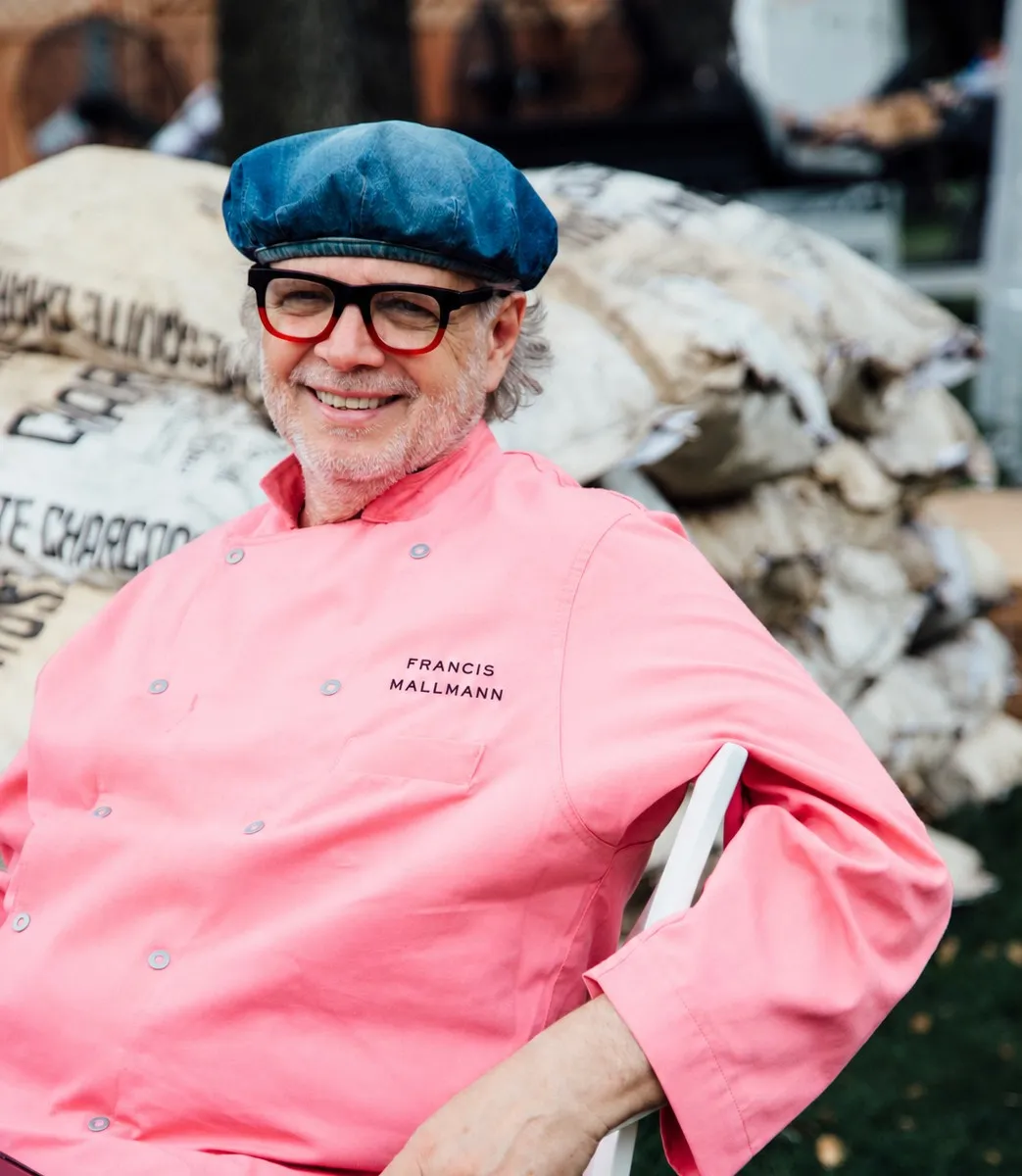 Francis Mallmann smiling in a pink chef jacket embroidered with his name, seated in front of charcoal sacks
